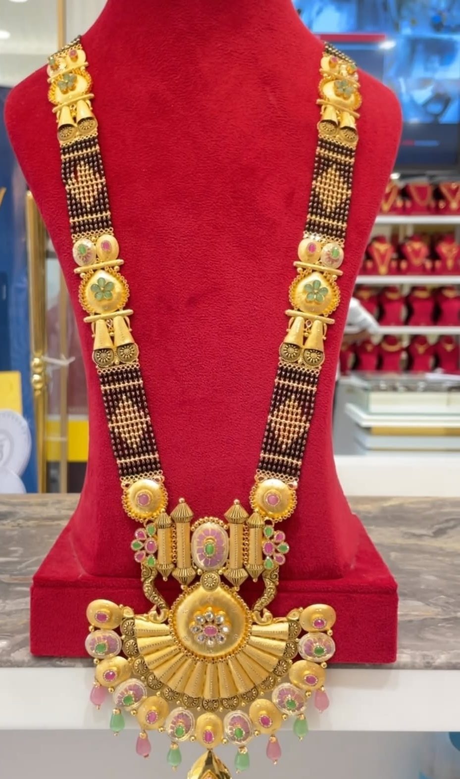 A long 22K gold mangalsutra with black beads and coloured stone drops on a red velvet stand